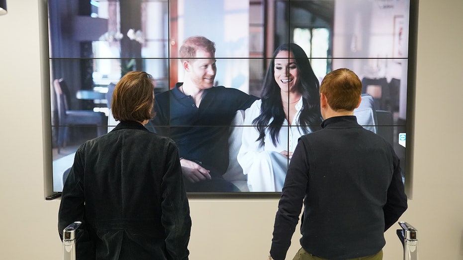 Two office workers with their backs turned watching Meghan and Harry on TV