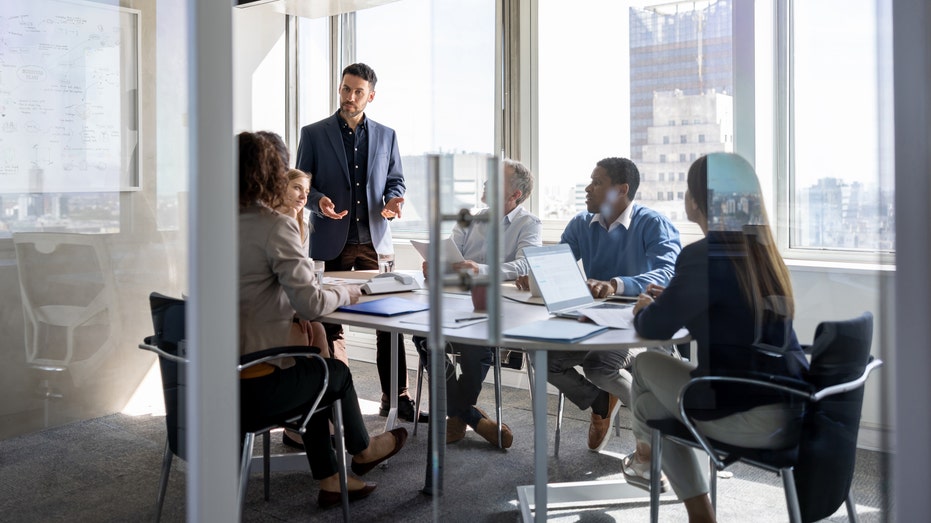 man standing talking to sitting colleagues at a table in an office